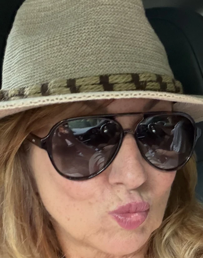 Sandy blowing a kiss in a sun hat and sunglasses